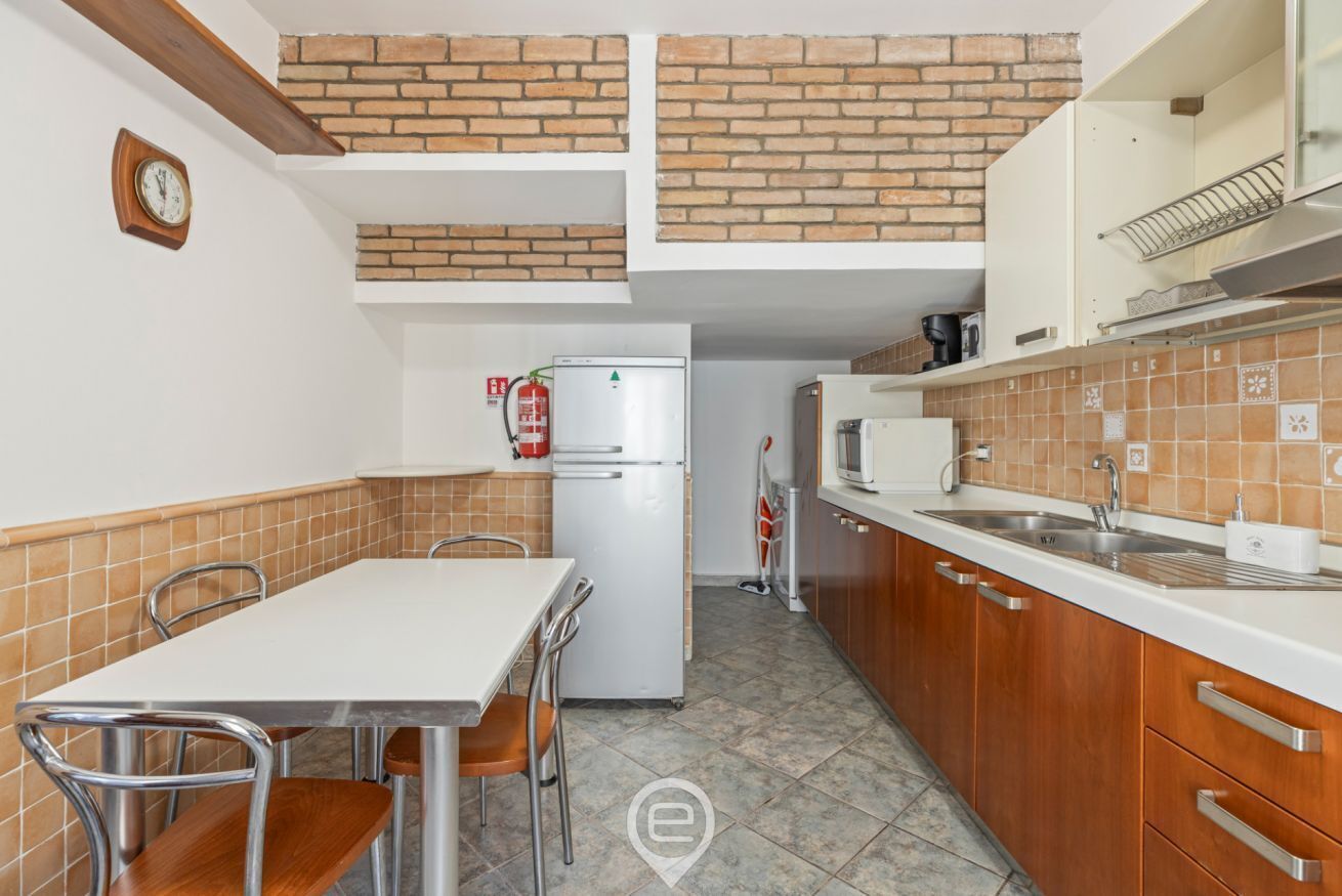 Eine kompakte Küche mit einem weißen Kühlschrank, einer Mikrowelle und einem Spülbecken. Ein Esstisch mit Stühlen und eine Uhr an der Wand sind ebenfalls vorhanden.