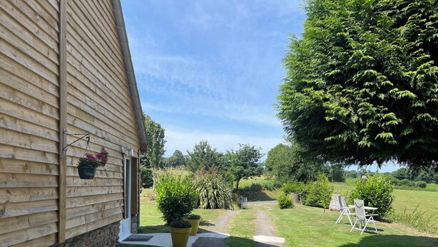 A narrow pathway leading to a building with a stone foundation and wooden siding. There are potted plants along the path, and a large tree provides shade to the right. - Charming 1-bedroom gite in delightful countryside hamlet with views (Saint-Georges-de-Livoye)