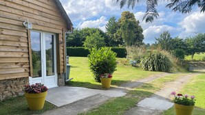 A wooden house with a stone foundation, a concrete pathway, and potted plants with flowers. - Charming 1-bedroom gite in delightful countryside hamlet with views (Saint-Georges-de-Livoye)