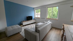 Un salon moderne avec un mur d’accent bleu, un ensemble de canapés, un téléviseur posé sur une console blanche et de grandes fenêtres donnant sur un paysage verdoyant.