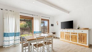 A dining area with a wooden table and chairs, a TV on a white cabinet, and a view of the outdoors through large windows.