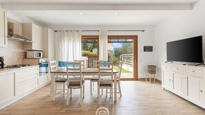 A modern kitchen with white cabinets, a dining area with a table and chairs, a TV on a cabinet, and a view of a garden through a glass door. - Garden Delight House (Chia)