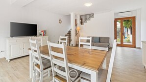 A modern living room with a wooden dining table, white chairs, a sofa, a TV, and a staircase. - Garden Delight House (Chia)