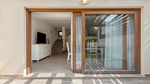 A modern house entrance with a wooden door and glass sliding doors, leading to a staircase and a living area with a TV and cabinets. - Garden Delight House (Chia)