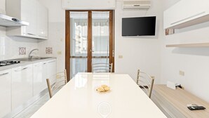 Una cocina moderna con armarios blancos, una mesa de comedor con sillas, un televisor montado en la pared y un equipo de aire acondicionado.