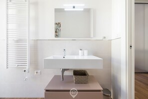 Un baño moderno con un lavamanos blanco, un espejo encima y un pequeño cubo de basura debajo.