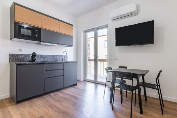 Una cocina moderna con armarios grises, un microondas negro y un horno empotrado. Hay una zona de comedor con una mesa y sillas, y un televisor de pantalla plana grande montado en la pared.