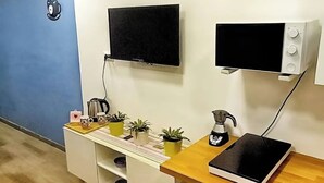 A compact kitchen with a microwave, kettle, and a small refrigerator. There are potted plants on the counter and a blue wall in the background.