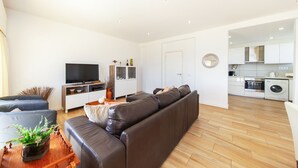 A modern living room with a brown leather sofa, a flat-screen TV, and a kitchen area with white cabinets and appliances. - Penthouse San Lorenzo (Portimão)