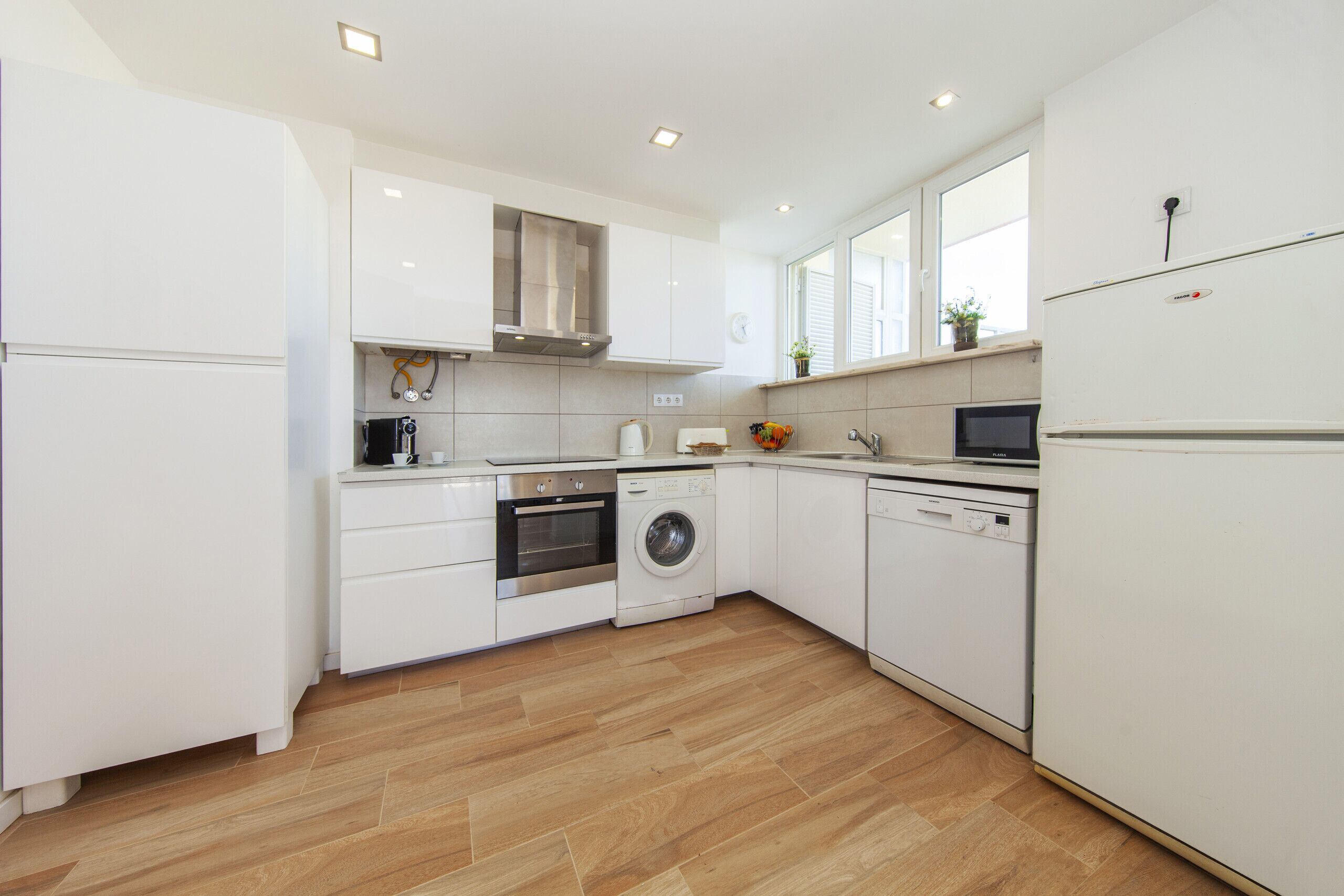 A modern kitchen with white cabinets, a stainless steel range hood, a washing machine, and a dishwasher.