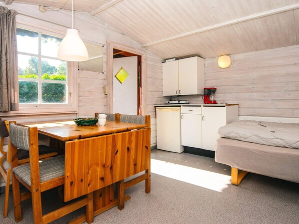 A compact room with a wooden dining table, a single bed, a kitchenette with a red coffee maker, and a window with curtains.