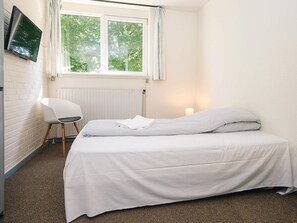 A simple, clean room with a bed, a chair, a TV mounted on the wall, and a window with curtains.