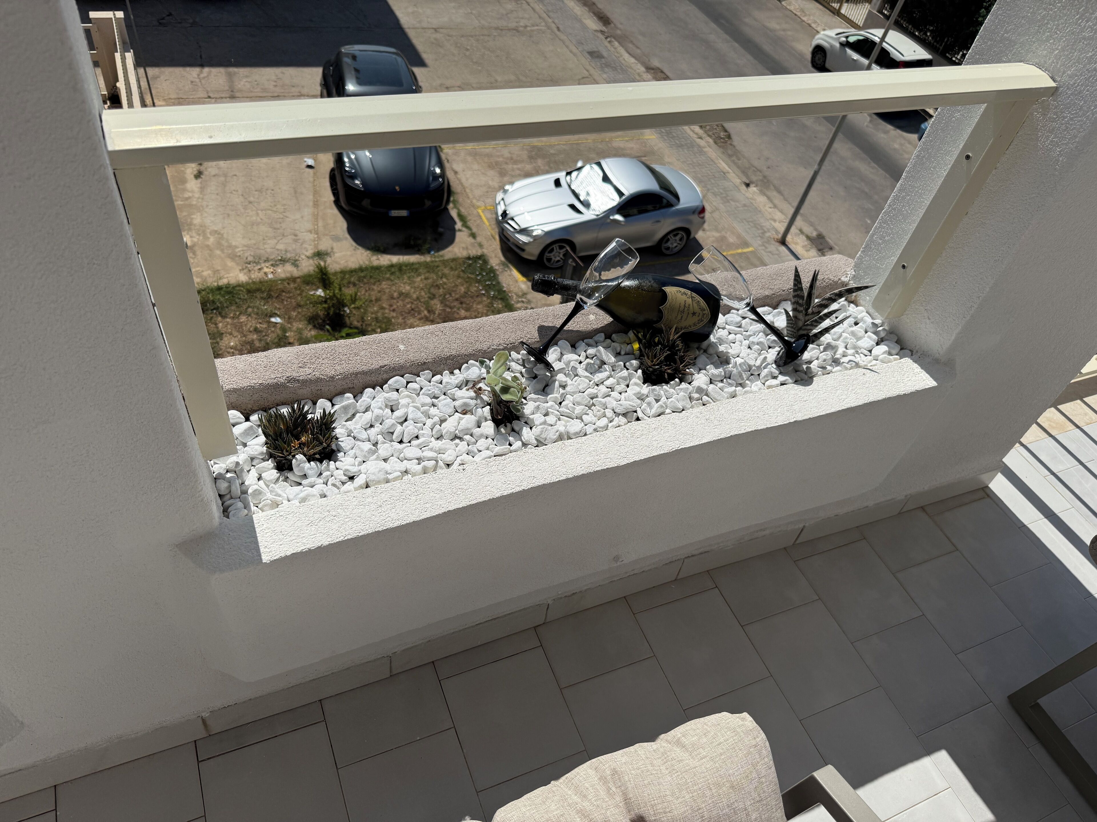 A balcony with a white railing, a small planter with white pebbles and plants, and a view of parked cars on the street below.