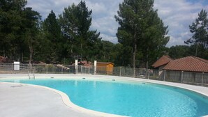 A swimming pool with a curved edge, surrounded by a fence, trees, and buildings with red-tiled roofs. - Holiday Home Under the Pines with Shared Pool, Private Terrace, and Wi-Fi (Moliets-Et-Maa)