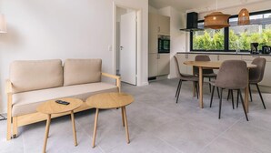 A modern living room with a sofa, two round wooden tables, and a dining area with chairs and a table. There is a kitchen visible through an open door. - 4 Person Holiday Park Home in Gilleleje (Gilleleje)