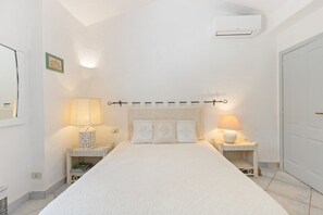 A bedroom with a large bed, two bedside tables with lamps, a wall-mounted air conditioner, and a door. - Villa Panta Rei - Liscia di Vacca (Liscia di Vacca)