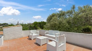 Un patio al aire libre con mobiliario blanco de mimbre, incluyendo un sofá y dos sillas, sobre un piso de baldosas.