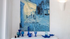 Una pintura de una escena callejera con edificios y gente, exhibida sobre una repisa con cristalería azul.