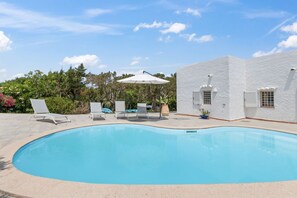 Piscina con sillones de descanso y sombrillas, rodeada por un edificio blanco con ventanas.