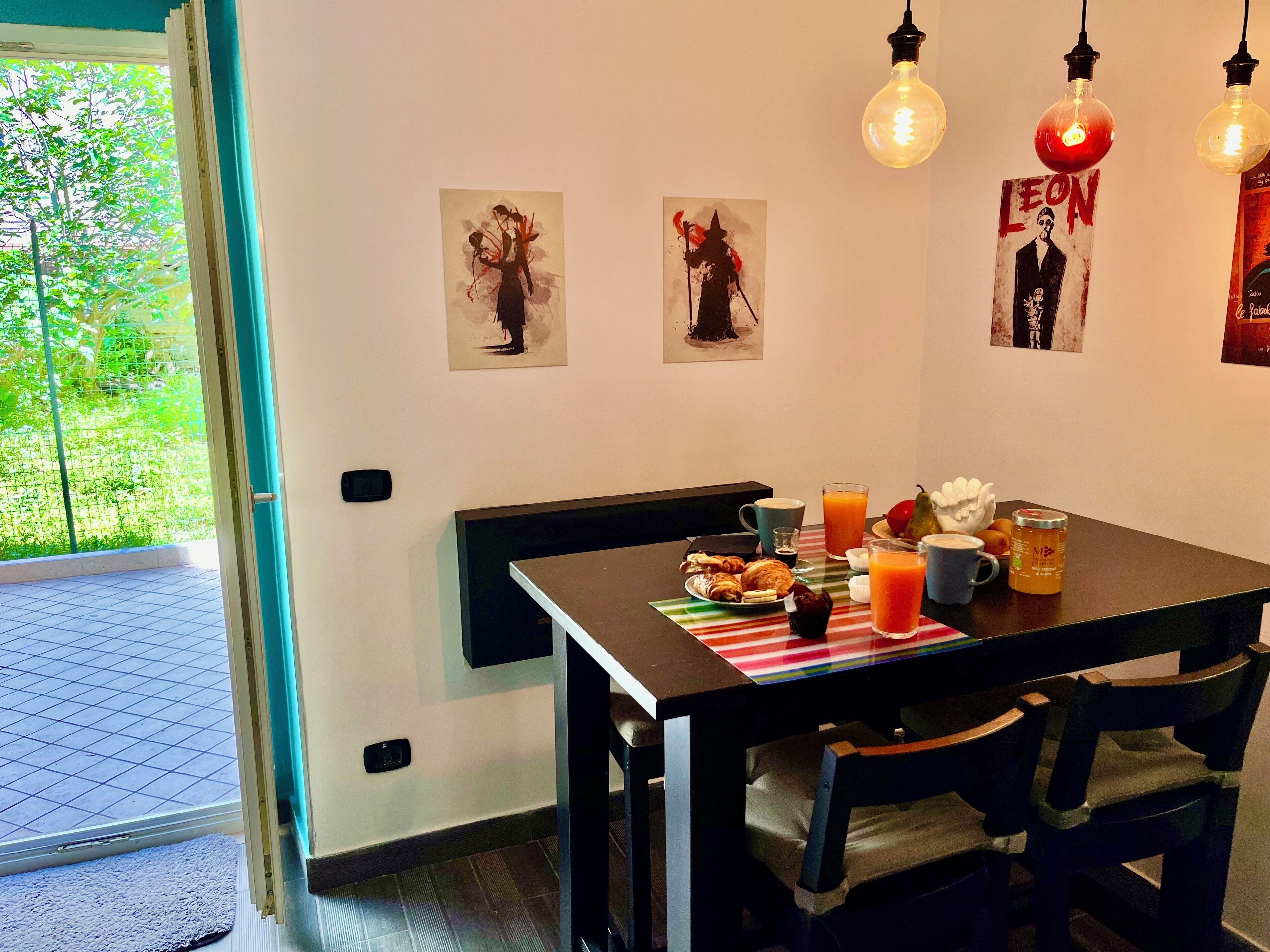 Zona pranzo con tavolo apparecchiato per la colazione, inclusi croissant, succo di frutta e una tazza di caffè. Tre lampade a parete e due opere d'arte incorniciate sopra il tavolo.