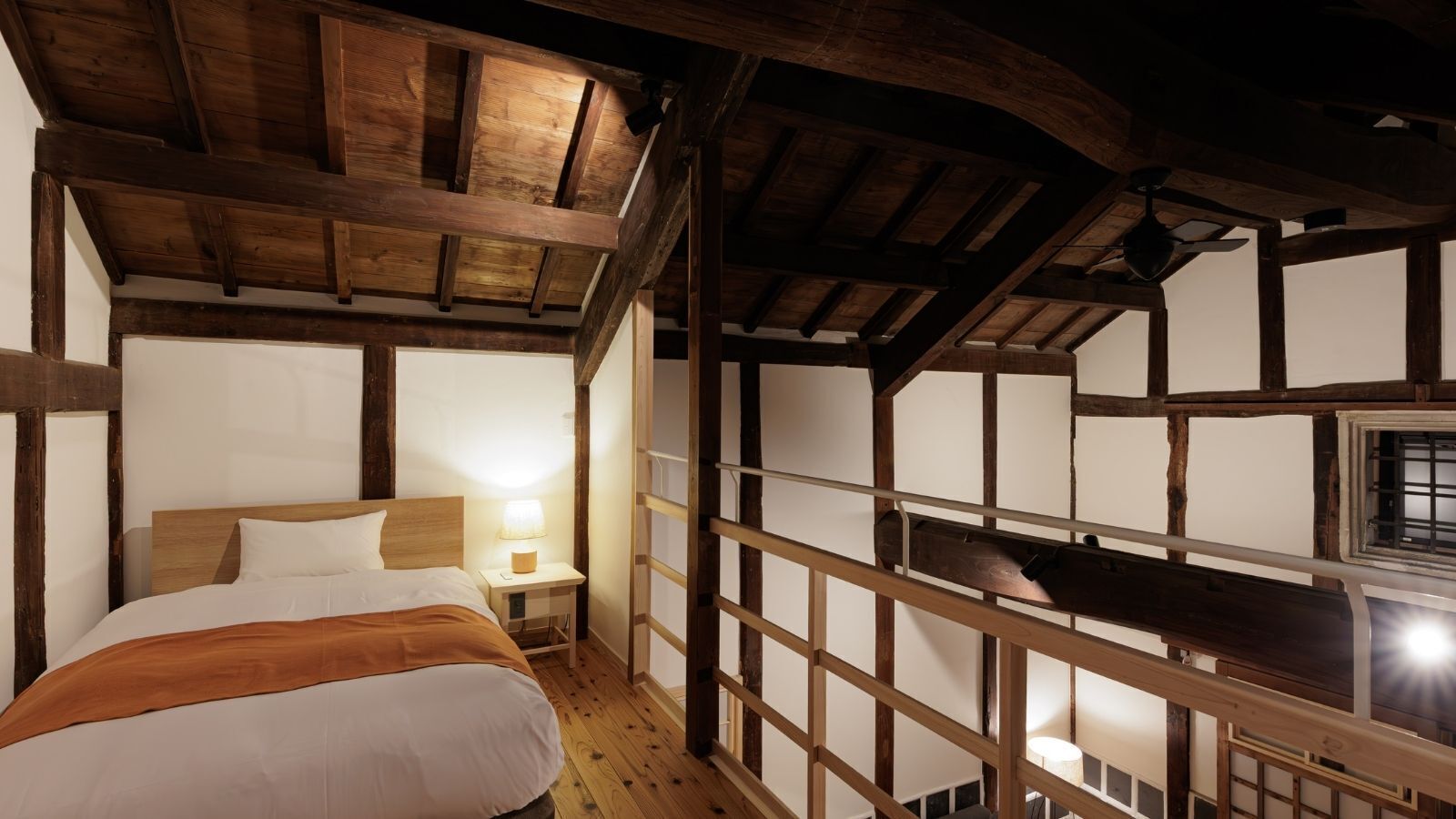 A bedroom with a wooden ceiling, a bed with white bedding, a nightstand with a lamp, and a window with a view.