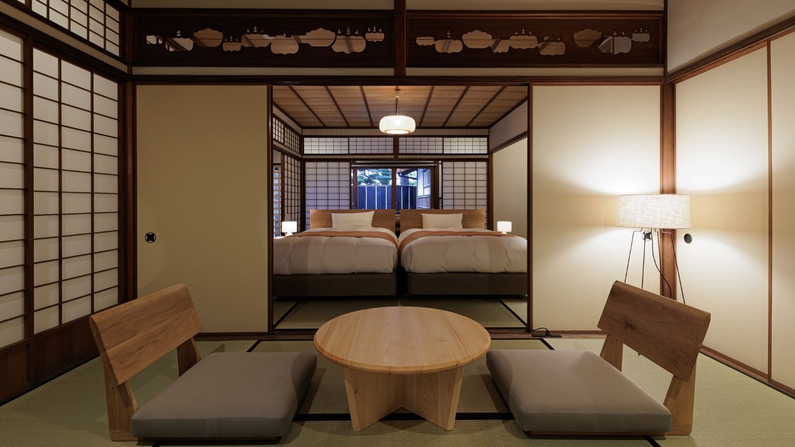A traditional Japanese room with tatami flooring, a round wooden table, two low wooden chairs, and a bed with a futon.