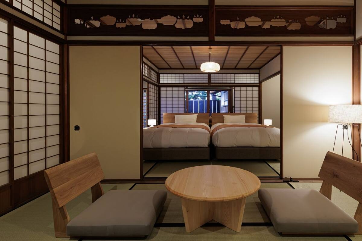 Une pièce de style japonais traditionnel, avec un sol recouvert de tatamis, une table ronde en bois, deux chaises basses en bois et un lit avec un futon.