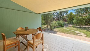 A covered patio with a wooden table and chairs, a green wall, and a view of a garden with trees and a fence.