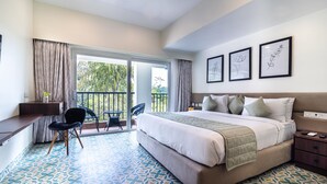 A hotel room with a large bed, a desk, a chair, and a balcony with a view of trees. - Ashreya Suites Tiruvanamalai (Tiruvannamalai)