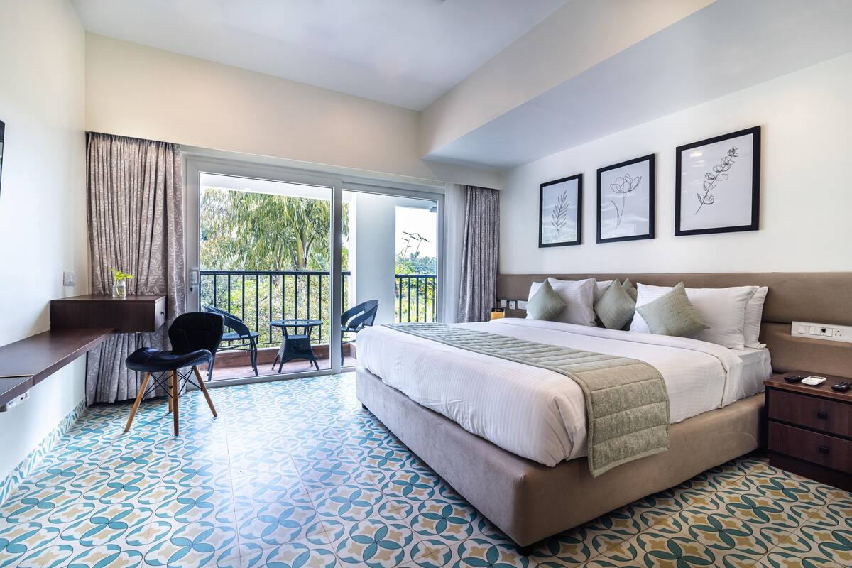 A hotel room with a large bed, a desk, a chair, and a balcony with a view of trees.