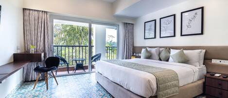 A hotel room with a large bed, a desk, a chair, and a balcony with a view of trees.
