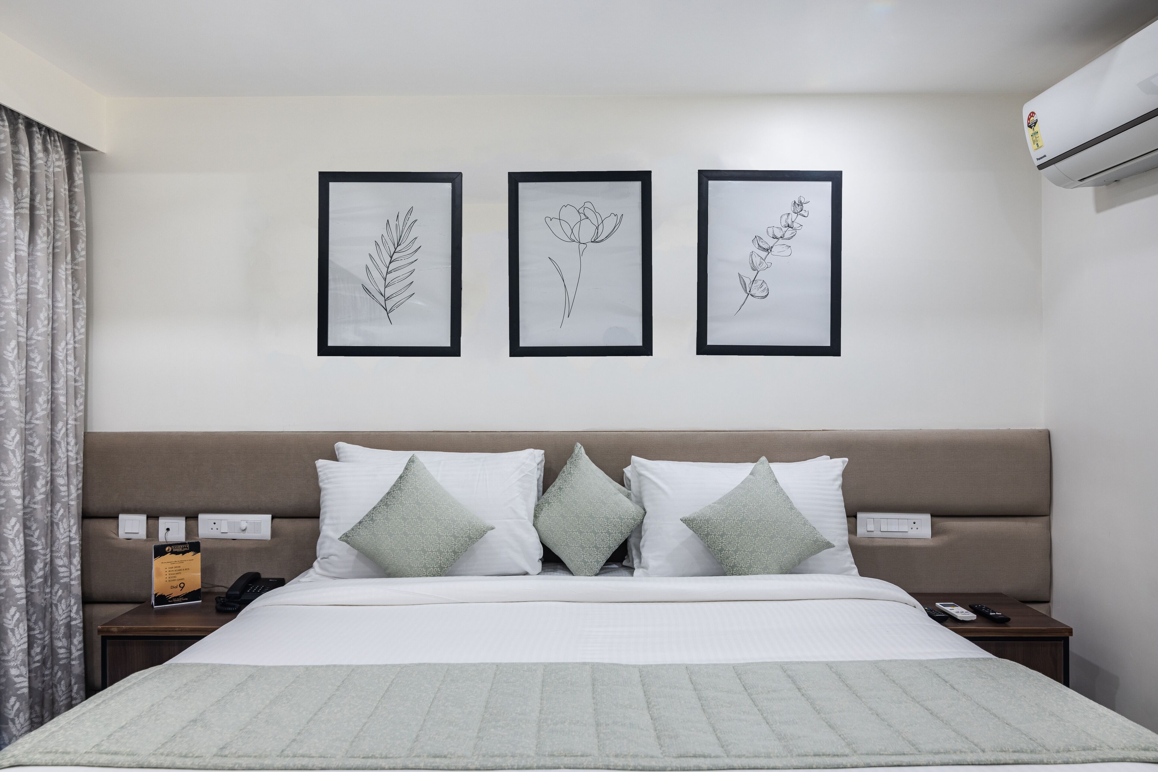 A neatly arranged hotel room with a bed, a nightstand, a wall-mounted air conditioner, and three framed botanical illustrations.
