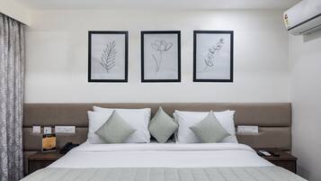 A neatly arranged hotel room with a bed, a nightstand, a wall-mounted air conditioner, and three framed botanical illustrations.