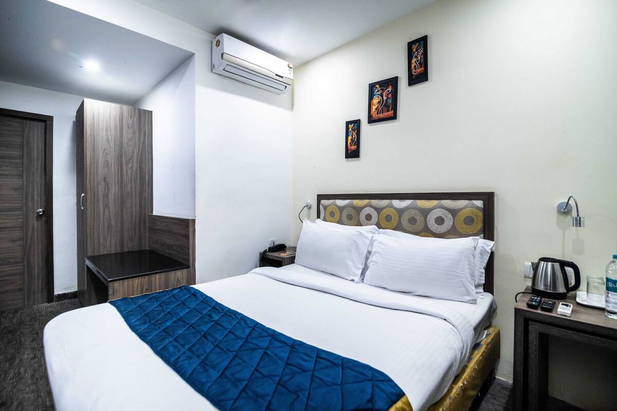 A hotel room with a bed, a nightstand, an air conditioner, a wall-mounted artwork, and a door.