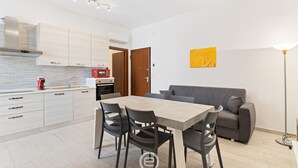 Une cuisine moderne avec des meubles blancs, une table à manger avec des chaises, un canapé gris et une œuvre d’art jaune accrochée au mur.