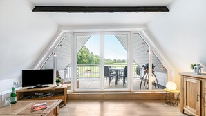 Ein geräumiges Dachzimmer mit einem großen Schiebeglasfenster, das auf einen Balkon führt, einer Holz-Unterhaltungseinheit und einem Essbereich mit Gartenmöbeln.
