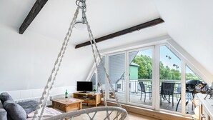 Een ruime kamer met een groot raam, een hangstoel, een houten tafel en een televisie.