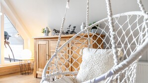 Een witte macramé hangstoel met een kussen, een houten kast en een klein tafeltje met een stoel in een lichte kamer met houten vloer.
