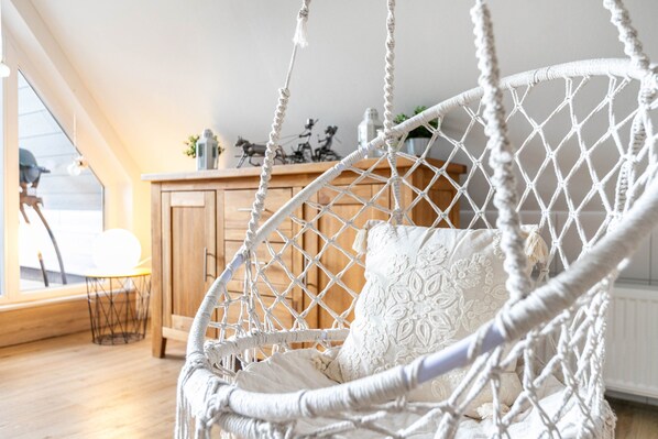 Una silla colgante de macramé blanco con un cojín, un aparador de madera y una mesita con una silla en una habitación luminosa con suelo de madera.