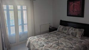 Une chambre à coucher avec un lit, un radiateur, une table de chevet, un tableau encadré et une fenêtre avec un rideau.
