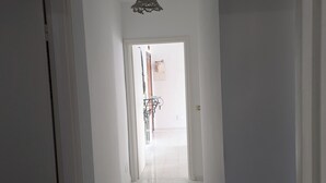 Un couloir étroit avec une porte blanche sur la gauche, un luminaire suspendu et, au fond, une vue sur une autre pièce où se trouve un vélo.