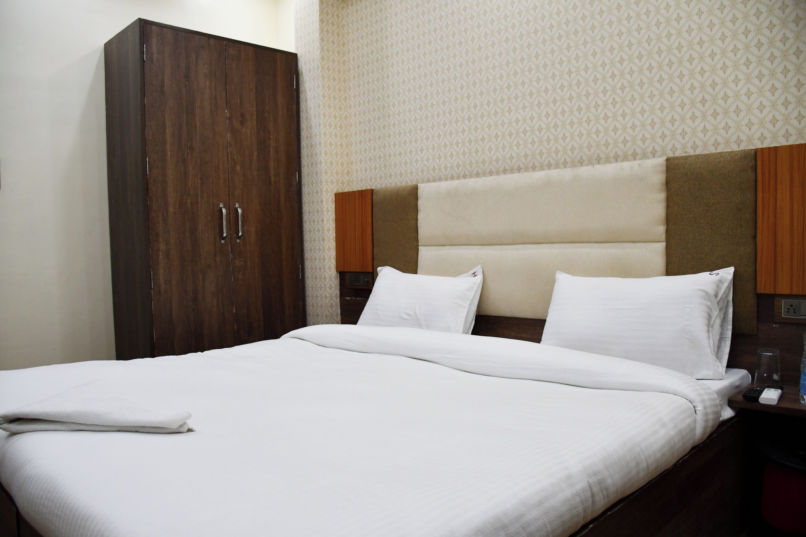 A neatly made bed with white linens, a wooden wardrobe, and a headboard with a brown panel.