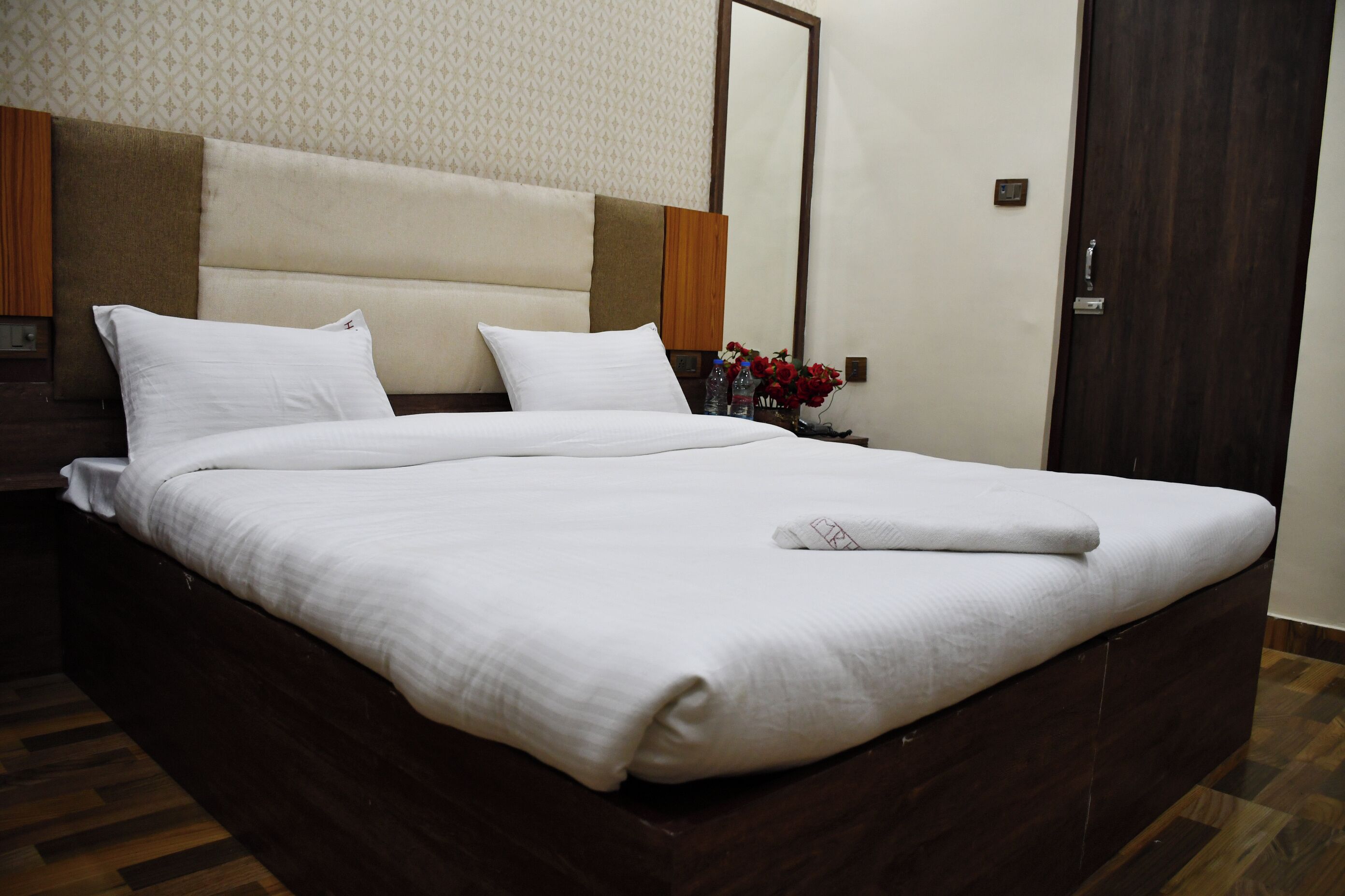 A neatly made bed with white linens, a wooden headboard, and a nightstand with a vase of flowers.