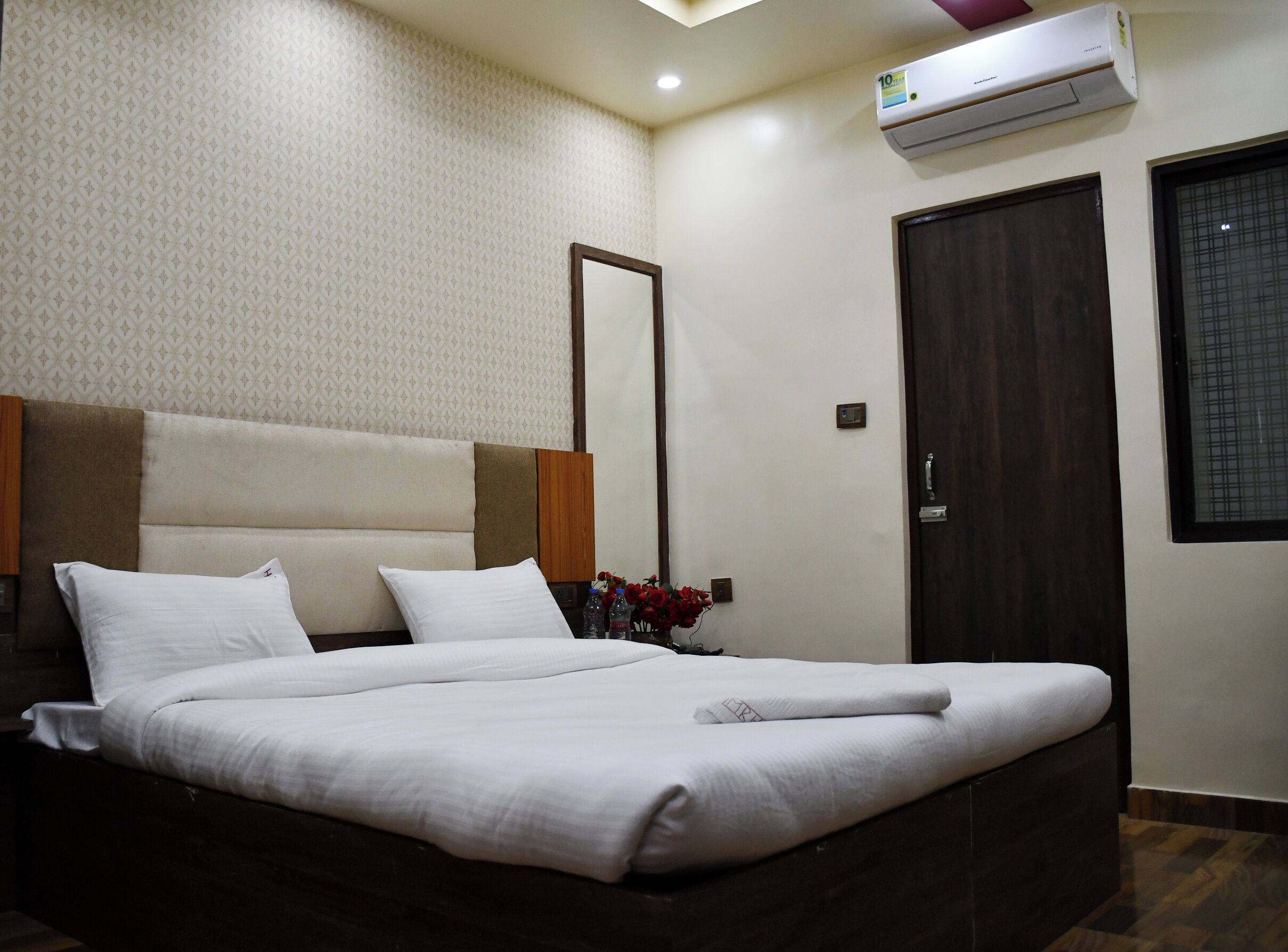 A neatly made bed with white linens, a headboard with a brown accent, and a wall-mounted air conditioner.