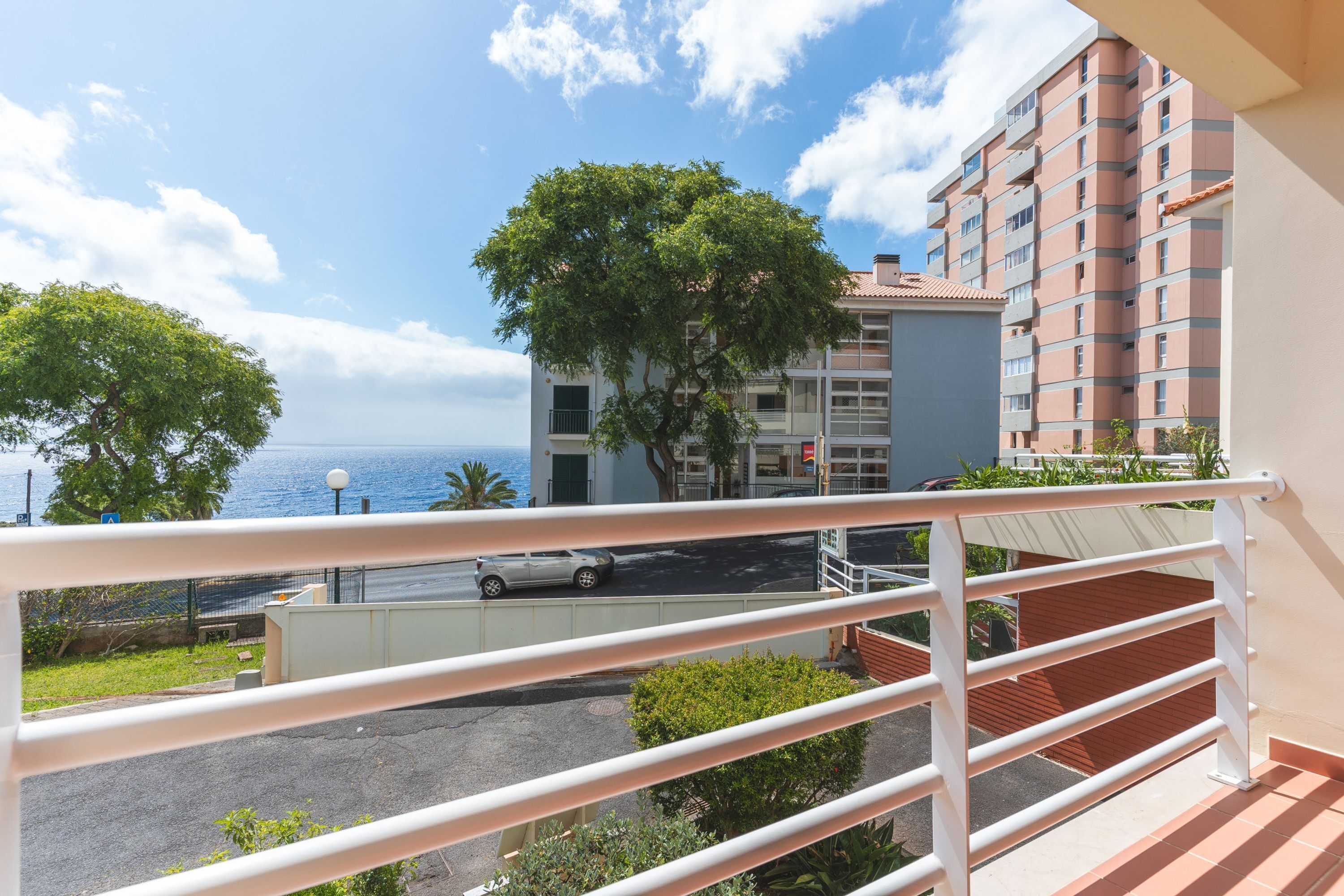 A balcony with a railing overlooking a parking area and a building with a red-tiled roof.