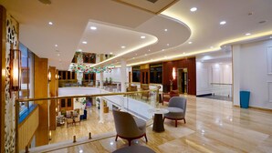 Un'ampia hall d'albergo con pavimento in marmo, ringhiera in vetro e una zona con sedute, poltrone e tavoli.