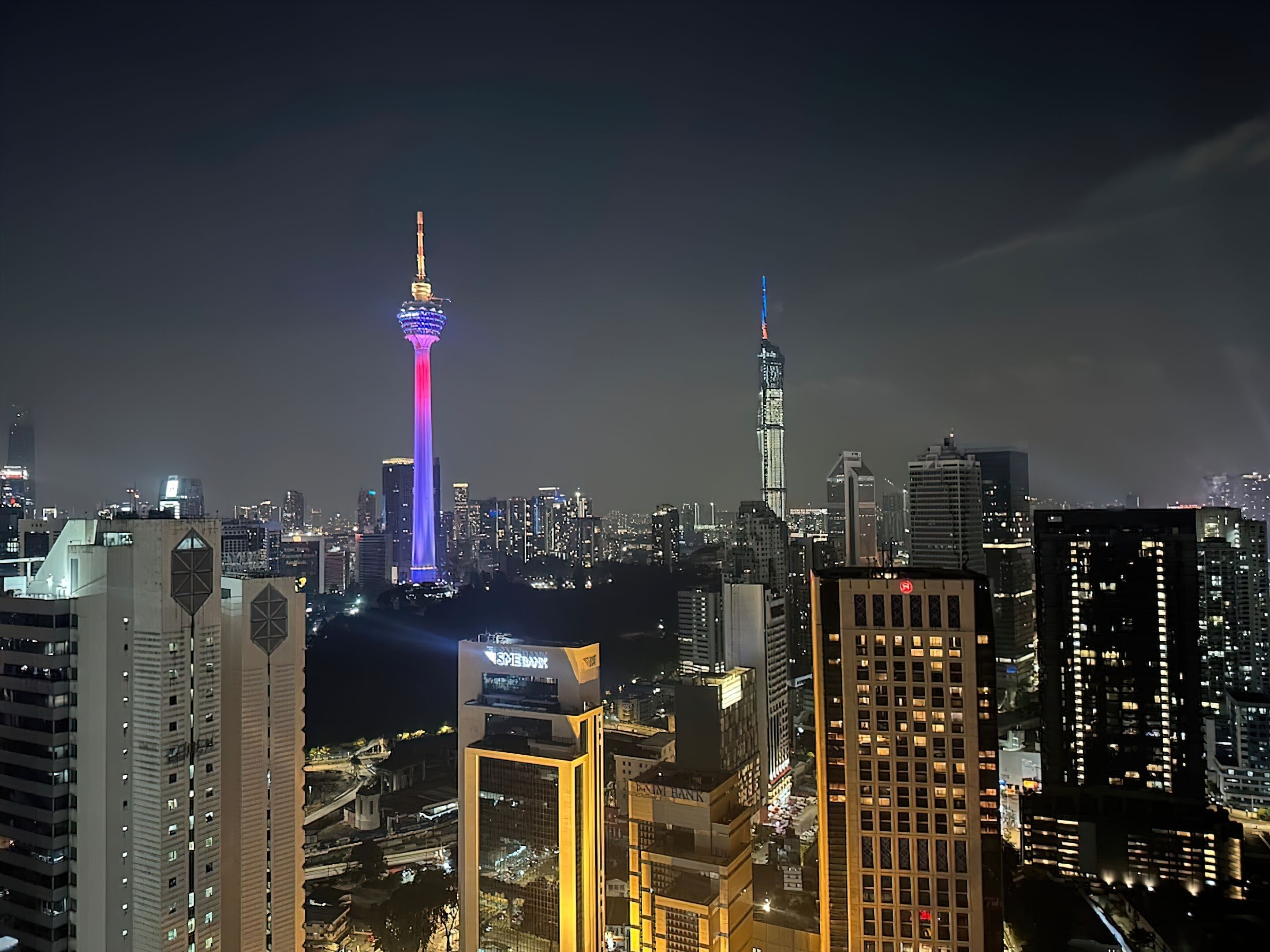 Uma paisagem urbana noturna com arranha-céus iluminados e uma torre imponente com um painel de luz vertical.