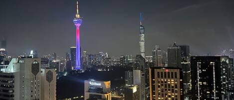 Uma paisagem urbana noturna com arranha-céus iluminados e uma torre imponente com um painel de luz vertical.