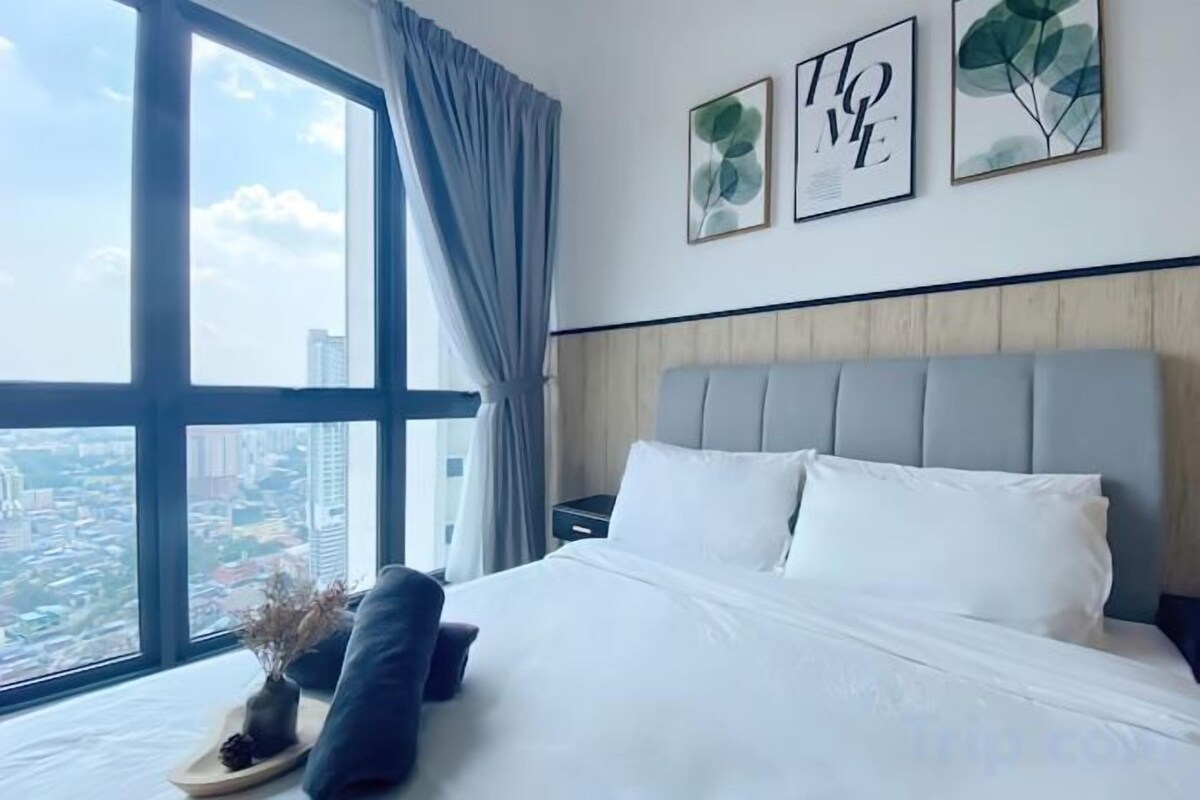 A hotel room with a large window, a bed with white linens, a grey headboard, and two framed artworks on the wall.