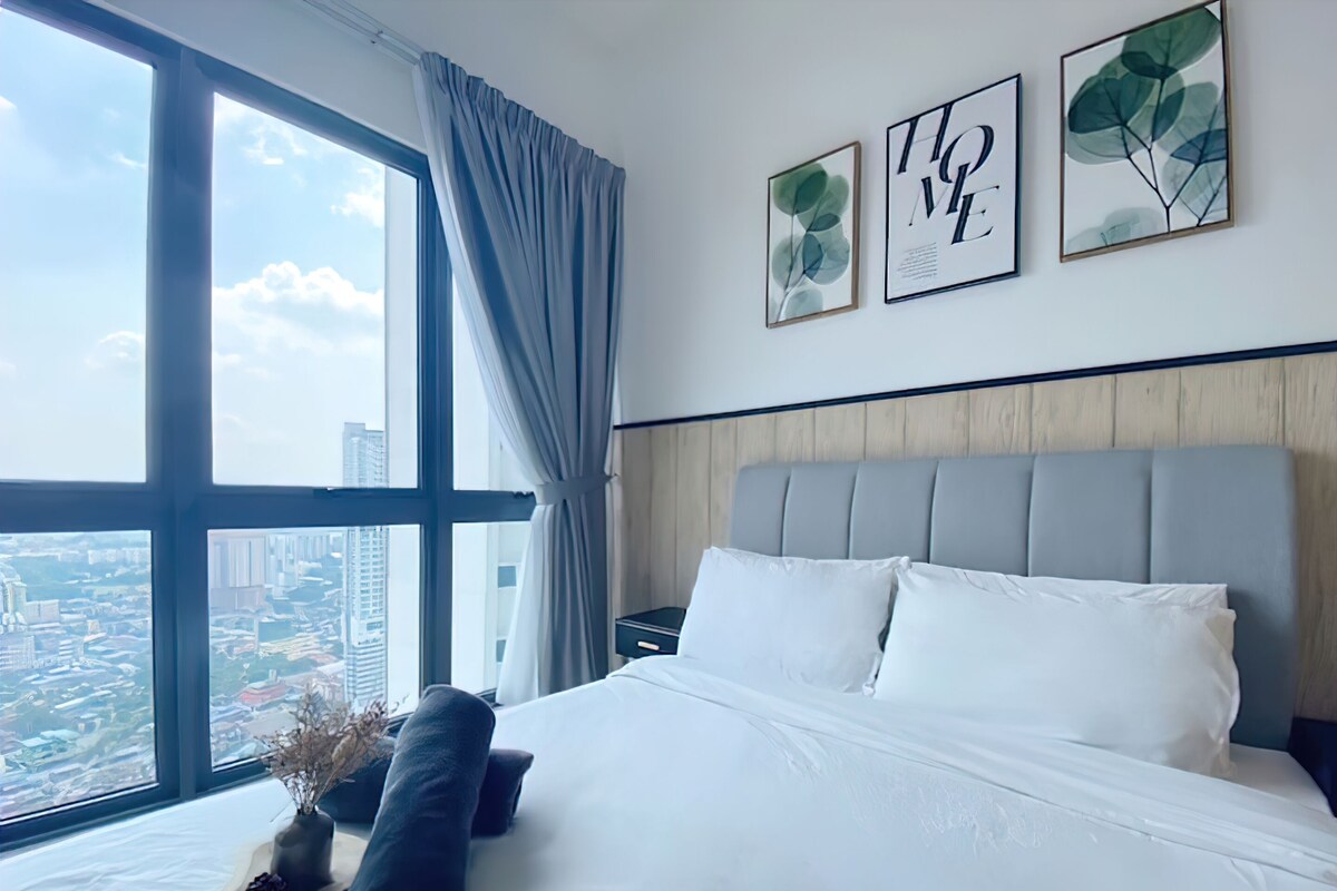 Habitación de hotel con un ventanal grande, una cama con ropa blanca, un cabecero gris y tres cuadros en la pared.